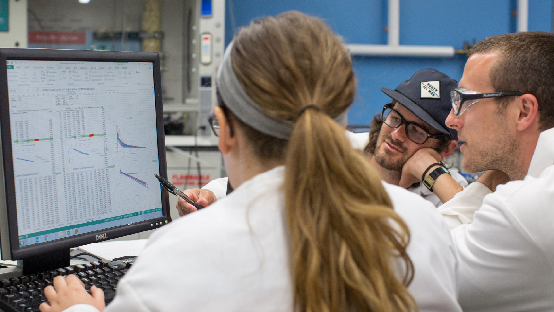 Three students in lab coats examine data on a computer